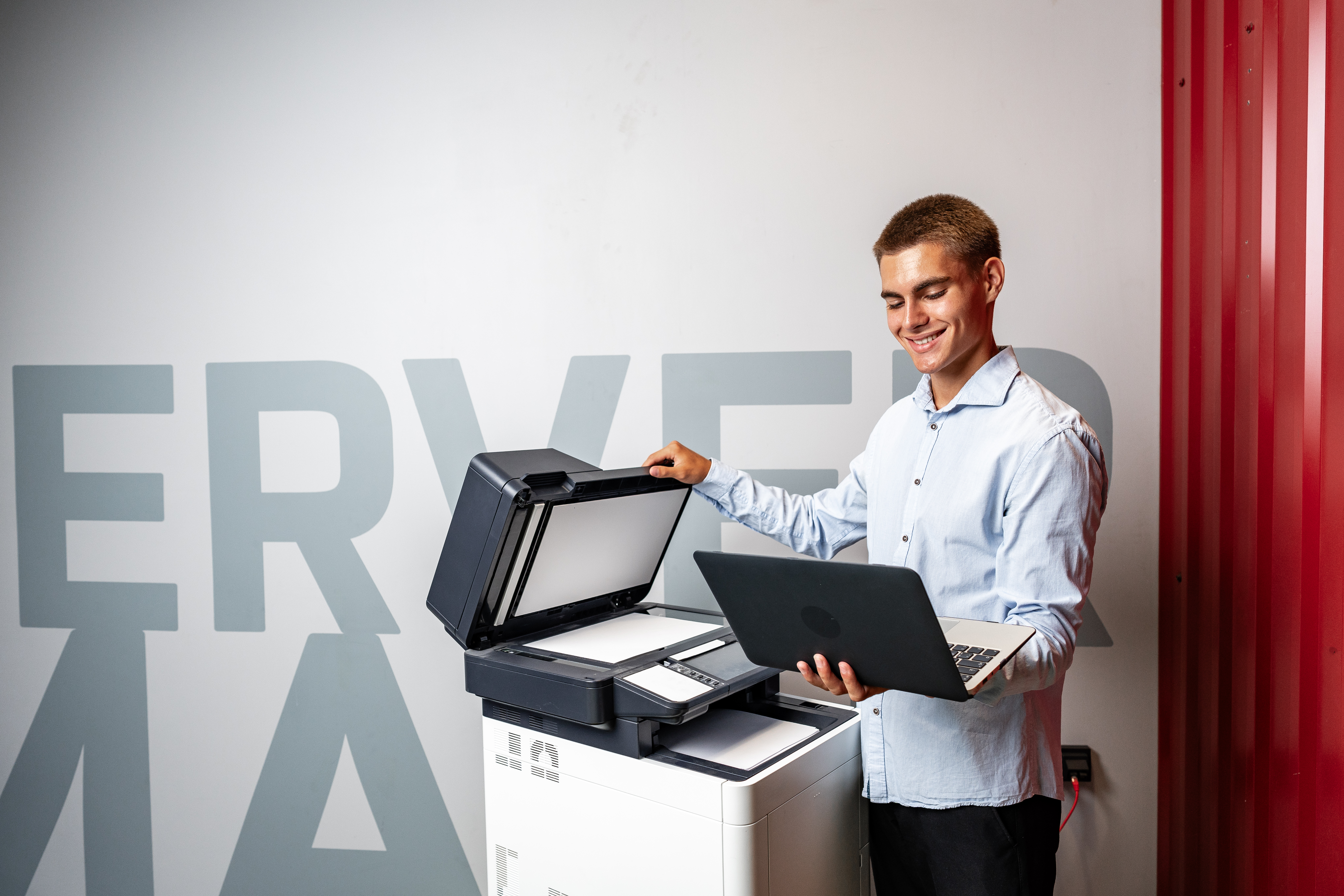 Small business employee using a laptop beside an office printer, showing how print management systems improve efficiency, control costs and simplify printing.